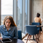 A young woman is seated by an office window with other employees in the background.